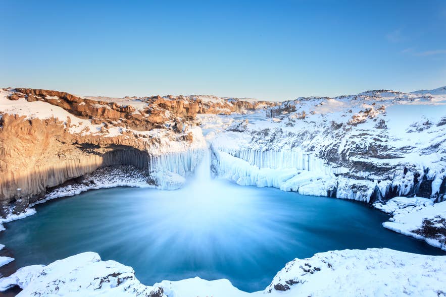 Aldeyjarfoss waterfall plunges into a snowy basalt basin in Iceland, glowing under a soft winter sunrise.