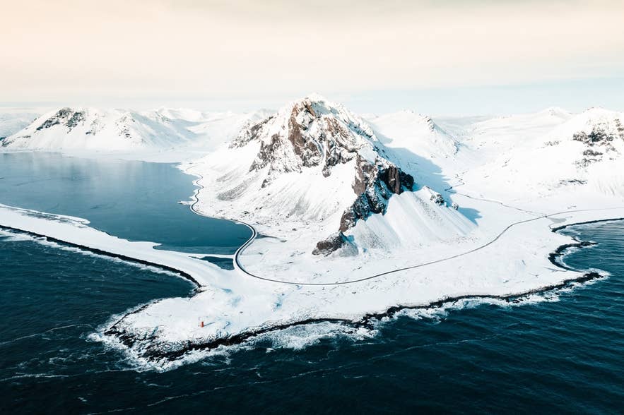 Der schneebedeckte Eystrahorn erhebt sich neben eisigen Gewässern in Ostisland, darunter eine kurvige Straße.