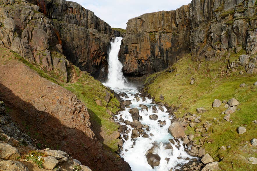 La cascata Fardagafoss scende tra pareti rocciose nell’Islanda orientale, circondata da pendii verdi e terreni aspri.