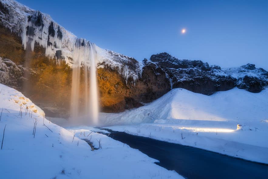 La cascata Seljalandsfoss brilla nel crepuscolo invernale, scorrendo accanto a stalattiti di ghiaccio in un paesaggio innevato sotto una luna crescente.