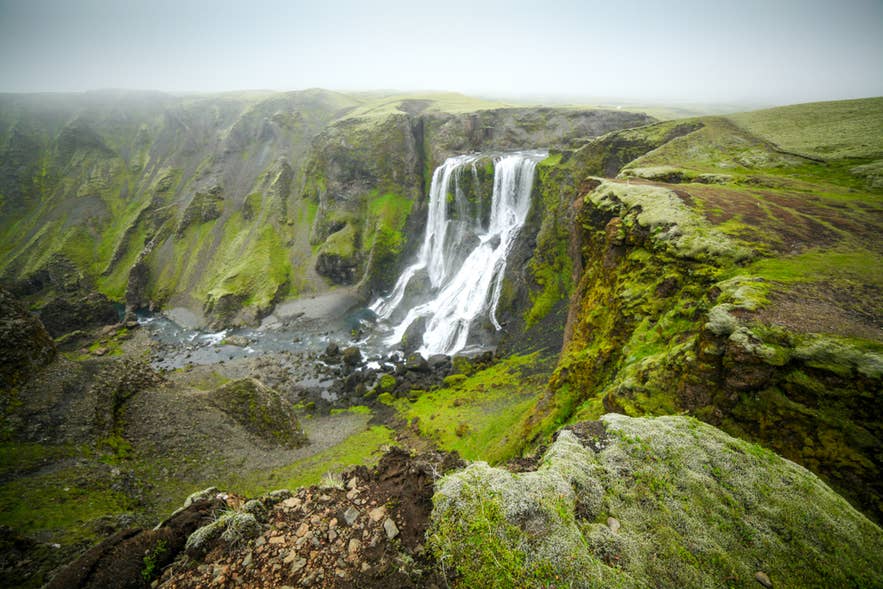 Der Ofaerufoss-Wasserfall stürzt durch eine moosige Schlucht in Island, eingerahmt von schroffen Klippen und nebliger Hochlandluft.
