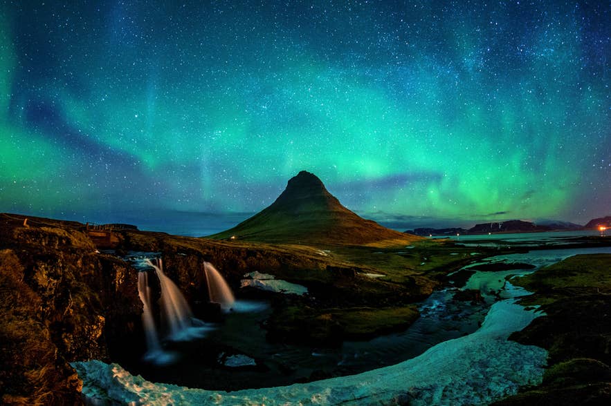 Der Kirkjufell leuchtet unter Nordlichtern in Island, mit den Kirkjufell-Wasserfällen im verschneiten Vordergrund.