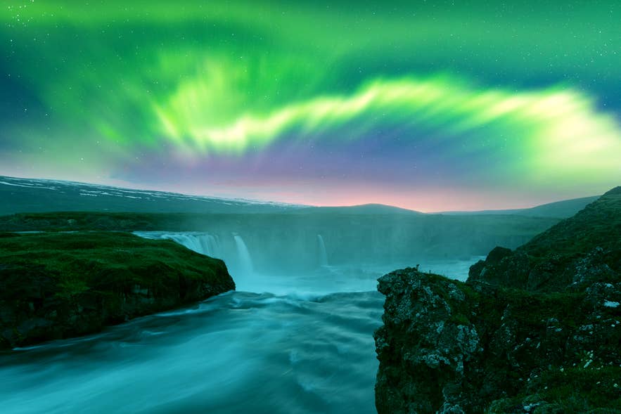 Der Godafoss-Wasserfall tost durch eine verschneite Schlucht in Nordisland, beleuchtet von leuchtend grünen und violetten Nordlichtern.