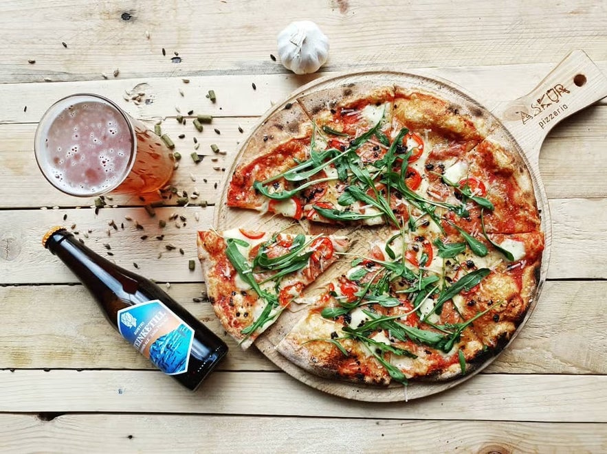 A wood-fired pizza topped with arugula, peppers, and melted cheese on a wooden board at Askur Pizzeria, served alongside a glass of beer and a bottled craft brew on a rustic wooden table.