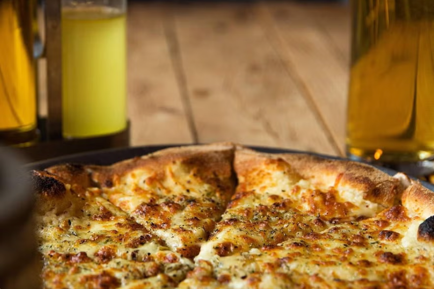 A close-up of a cheesy, golden-brown pizza at Askur Pizzeria, with glasses of beer and a yellow drink blurred in the background on a wooden table.