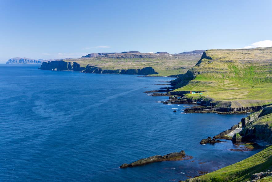 Een ruige groene kustlijn grenst aan diepblauw water in het Hornstrandir Natuurreservaat in de Westfjorden.