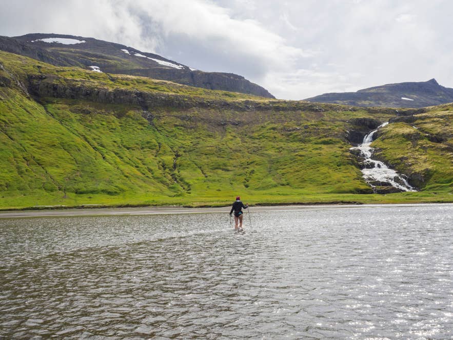 Een wandelaar waadt door ondiep water richting groene berghellingen en een waterval in het Hornstrandir Natuurreservaat.