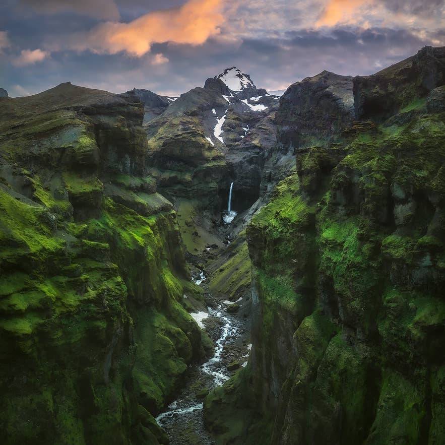 ムーラグリューフル渓谷の苔むした崖と細い川、遠くに滝と雪を頂く山。