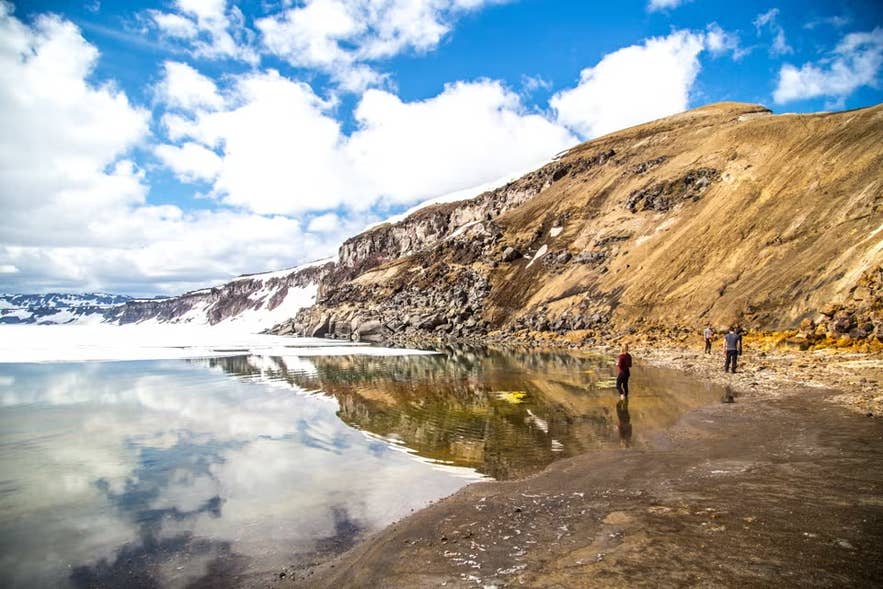Wandelaars verkennen de oever van het Askja calderameer, dat besneeuwde bergen weerspiegelt onder heldere wolken in Noord-IJsland.