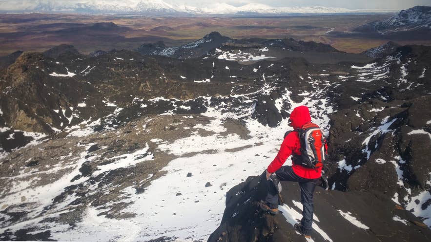 赤いジャケットを着たハイカーが、アイスランドのハフナルフィヨルズル近郊で雪に覆われた溶岩原を見下ろす黒い火山岩の上に立っている。