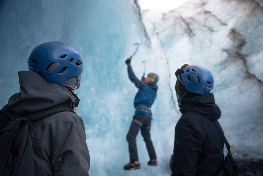Two ice climbers in helmets swinging an axe into a blue ice wall on a glacier in Iceland