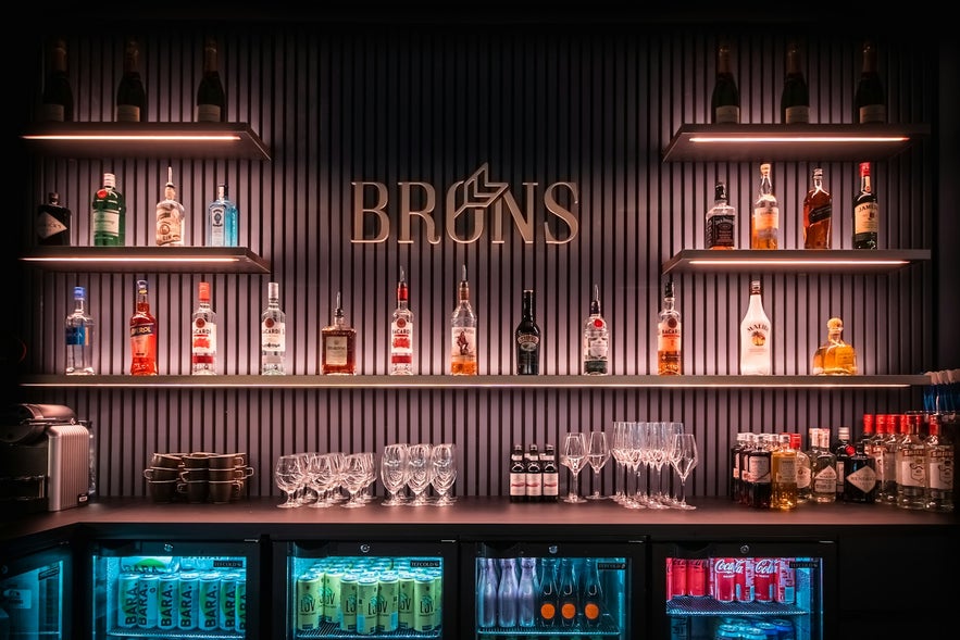 Well-lit Brons bar display with liquor bottles, glassware, and fridges stocked with drinks against a striped wall.