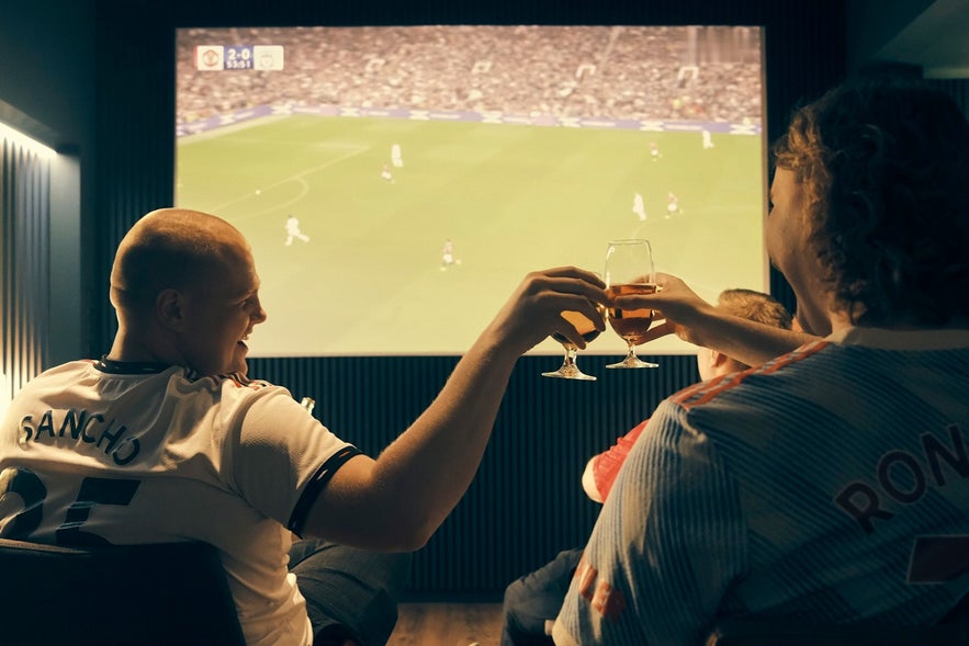 Brons Keflavik patrons cheer during a soccer match and toasting a drink in front of a screen.