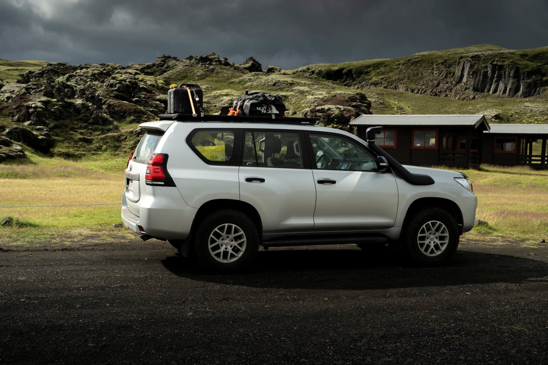 A white 4x4 Super Jeep with gear on the roof rack is parked on a grassy plain beside rocky hills under dark clouds in Iceland.