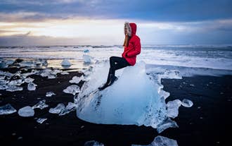 Kobieta w czerwonym zimowym stroju siedzi na ogromnym kawałku lodu na Diamentowej Plaży na południowym wybrzeżu Islandii.