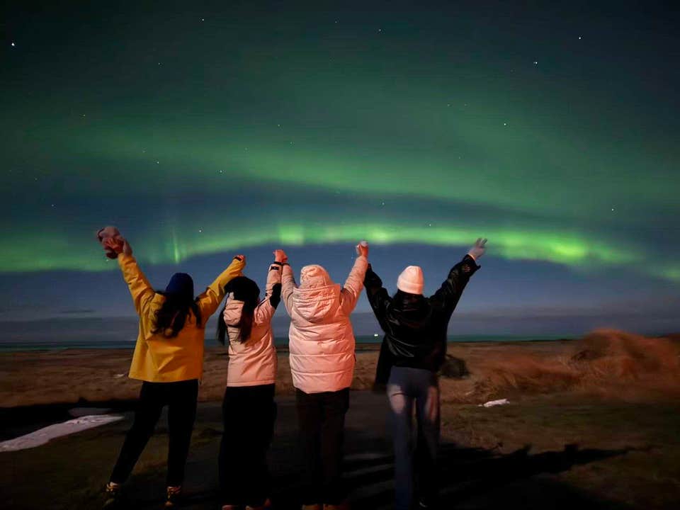 Viajeros celebrando bajo un brillante arco verde de auroras boreales durante una parada para ver la aurora en Islandia.