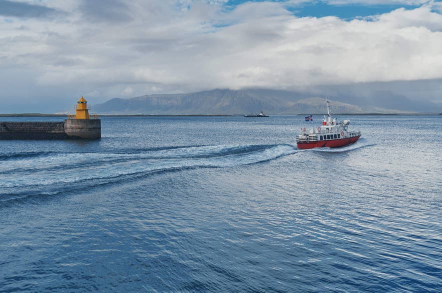 A whale-watching boat departs Reykjavik harbor, passing a lighthouse with mountains visible across Faxafloi Bay.