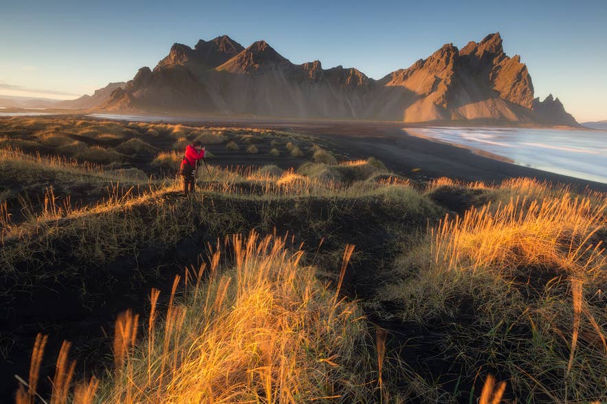 A photographer captures Vestrahorn at sunrise as golden light sweeps across black sand dunes and coastal grass in southeast Iceland.