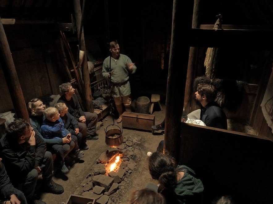 游客聚集在Eiríksstaðir的一座维京长屋内，围坐在敞开的炉火旁聆听导游讲解，重现历史场景。
