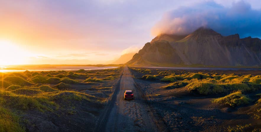Ein Auto fährt bei Sonnenuntergang auf einer abgelegenen Schotterstraße in Island, umgeben von moosbedeckten Lavafeldern und dramatischen Bergen.