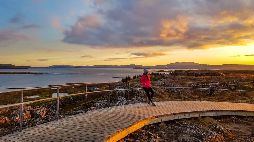 Person standing on a curved wooden viewing platform in Iceland at sunset, overlooking a lake, lava fields, and distant mountains.