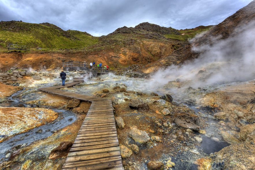 Visitors walk along wooden boardwalks through steaming vents and bubbling hot springs at Krysuv&iacute;k Geothermal Area in Iceland.