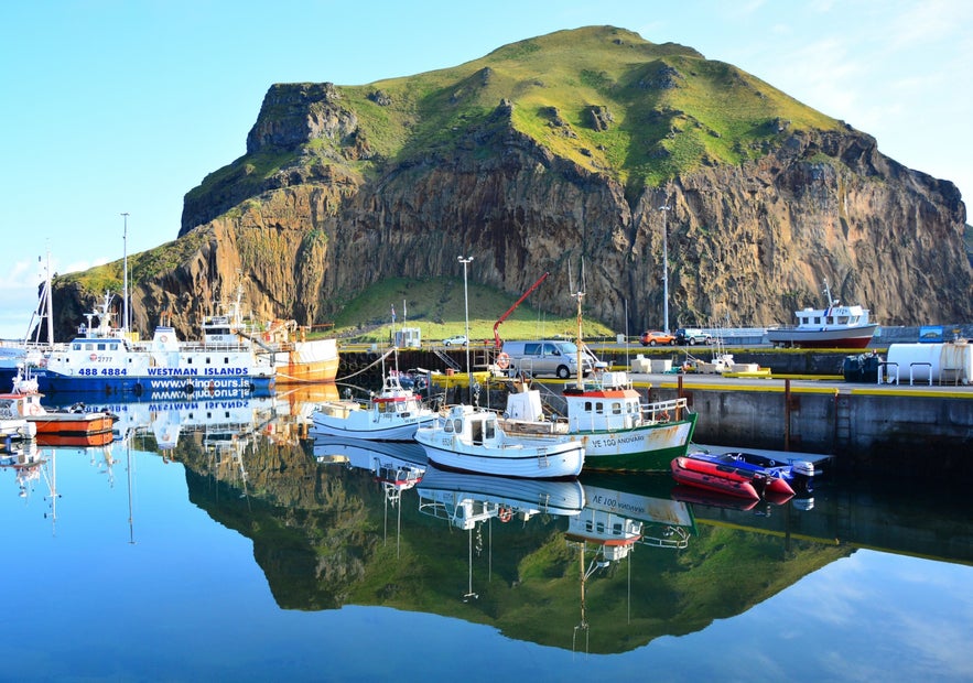 Heimaey Harbor in the Westman Islands, with fishing boats reflected below Heimaklettur Cliff in Vestmannaeyjar, Iceland.