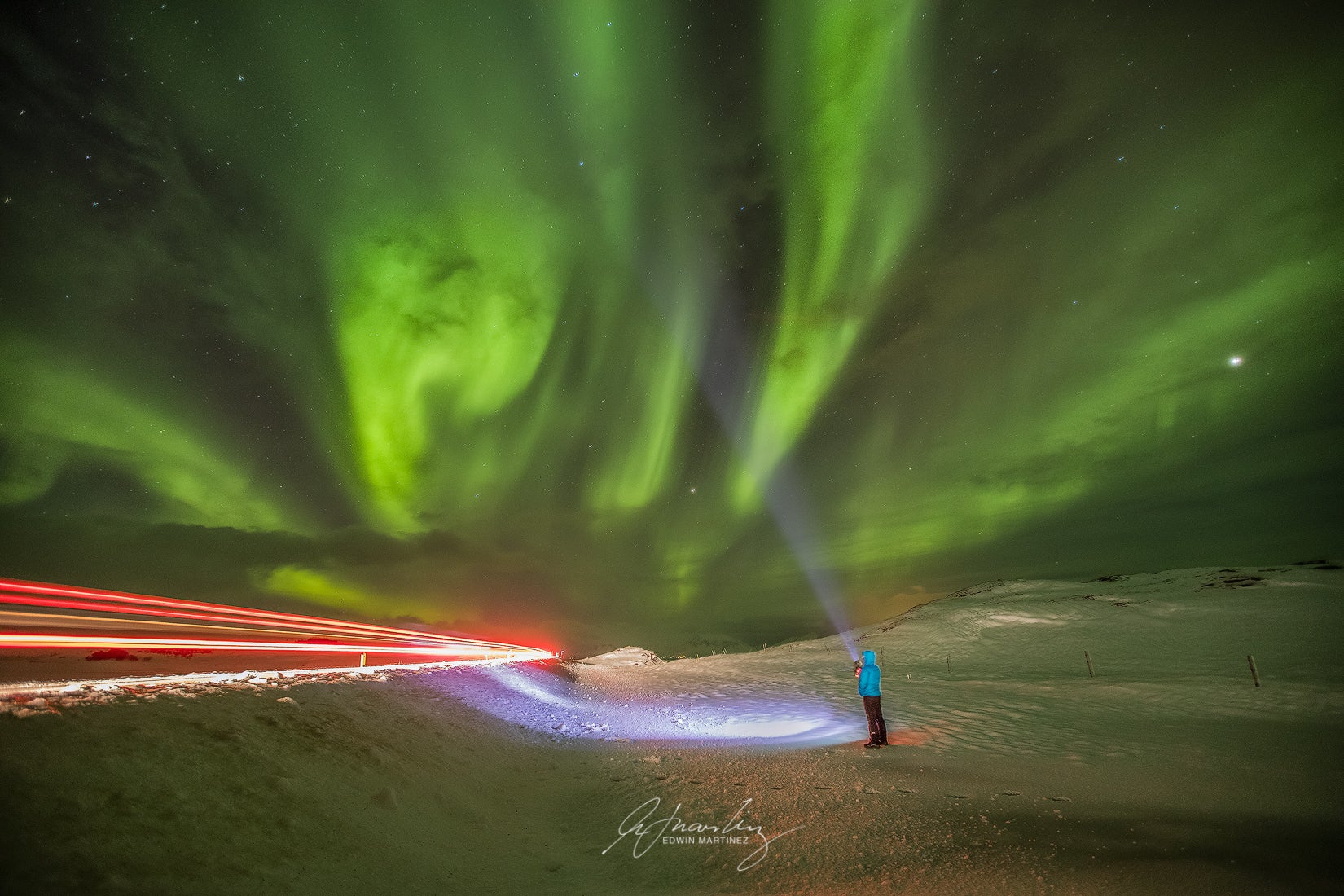 Traveler under vivid aurora borealis beams near snowy roadside in Iceland.