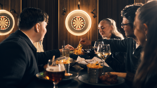 Friends sharing food and drinks at Brons Keflavik restaurant, enjoying Icelandic dining with dartboards and a lively atmosphere.