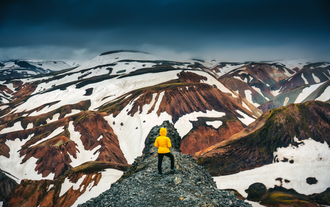 Hiker in yellow jacket standing on a rocky ridge overlooking colorful snow-covered mountains along the Laugavegur trail.