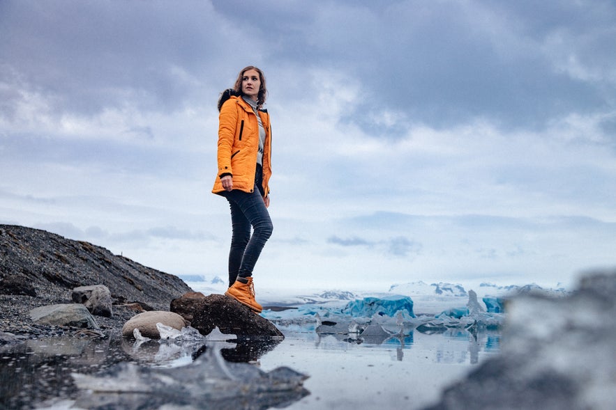 A traveler in a yellow jacket stands on a rock beside J&ouml;kulsarlon glacier lagoon, with floating ice and distant glaciers.