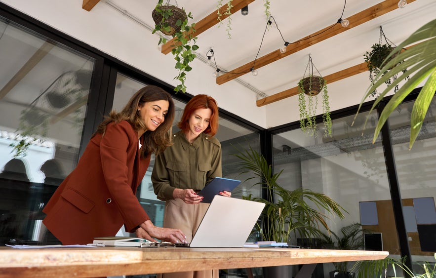 Two women collaborate at a desk with a laptop and tablet in a bright, plant-filled office space.