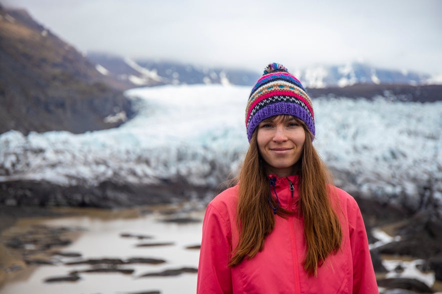 A traveler in a colorful Icelandic wool hat stands in front of a glacier and rocky landscape.