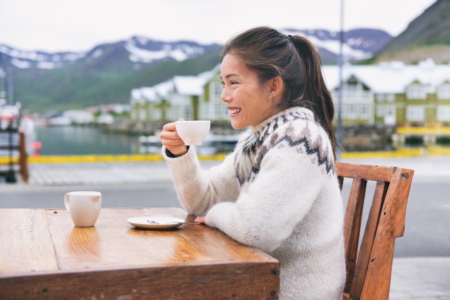 A traveler wearing an Icelandic wool sweater enjoys a cup of coffee at an outdoor table with a harbor and mountains in the background.