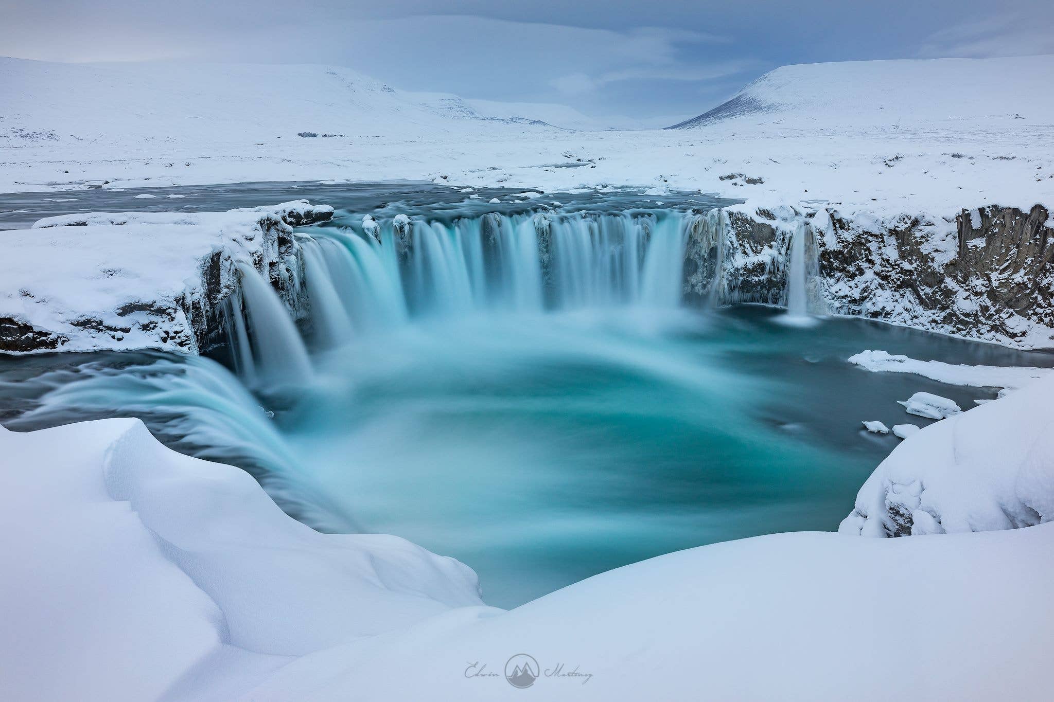 Snow-covered cliffs and turquoise water at Godafoss Waterfall in winter.