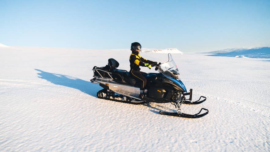 A person rides a snowmobile across Langjökull glacier in Iceland under a clear blue sky.