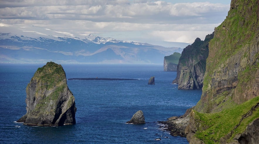 Dramatic sea cliffs and volcanic rock formations of the Westman Islands, Iceland, with views of Eyjafjallajokull glacier and the South Coast mountains in the background.
