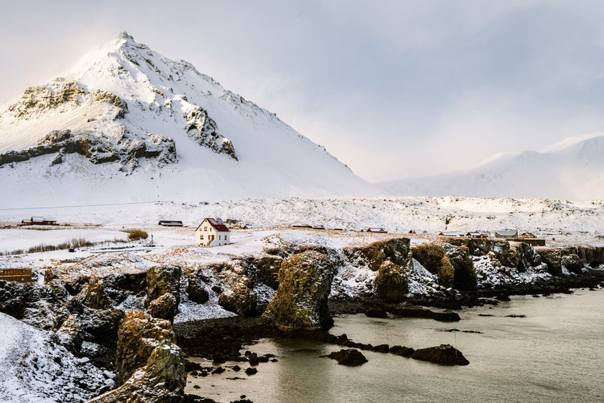 A lone house stands at the base of a snow-covered mountain in Arnarstapi, Iceland.