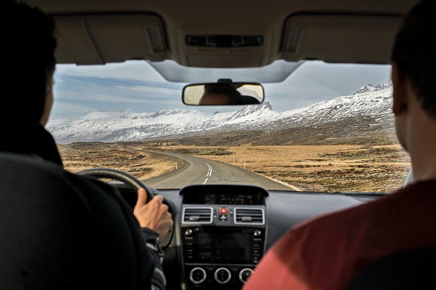 Two guys inside the car driving on the country roadway between fields with brown grass and snowy mountains on the cloudy sky background in Iceland. 