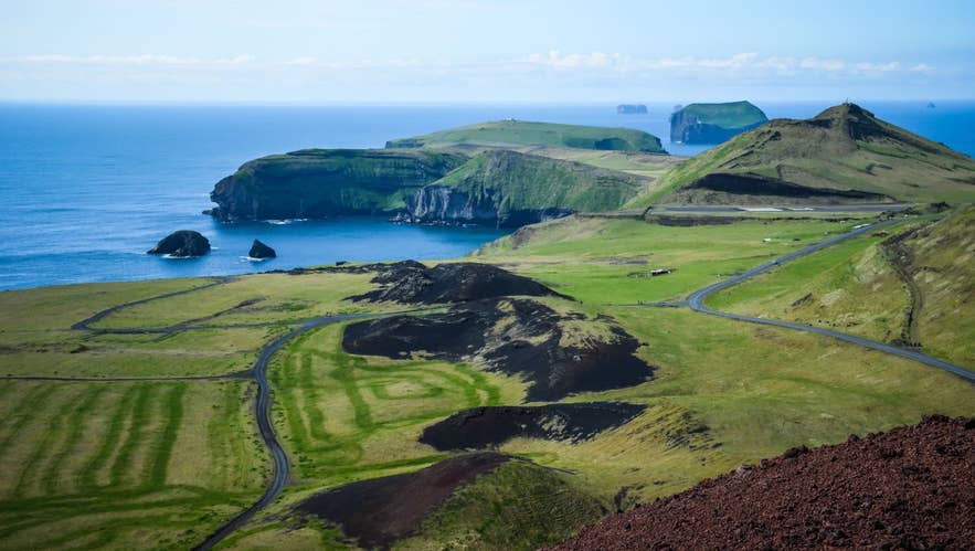 Panoramic view of Heimaey Island in the Westman Islands archipelago, Iceland, showing volcanic craters, green cliffs, and coastal sea stacks under a clear blue sky