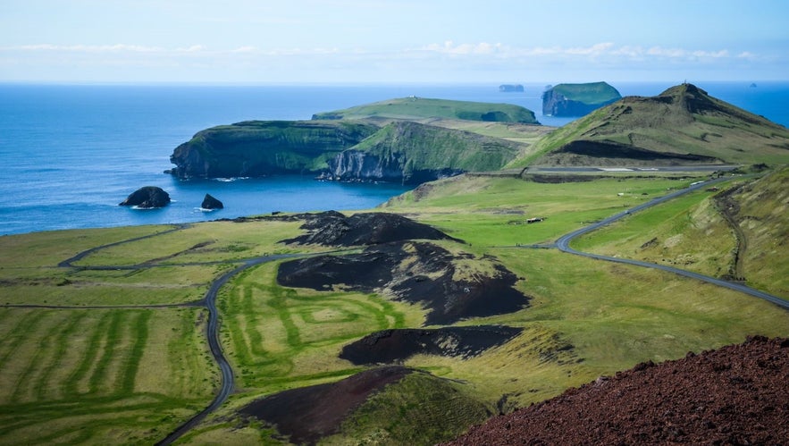 Panoramic view of Heimaey Island in the Westman Islands archipelago, Iceland, showing volcanic craters, green cliffs, and coastal sea stacks under a clear blue sky