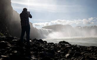 Man photographing the misty cascade of Godafoss Waterfall in North Iceland on a sunny day.