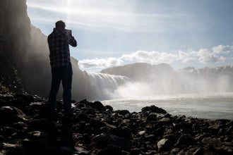Un fotógrafo captura la Cascada Godafoss desde un sendero rocoso junto al río bajo la luz del sol.
