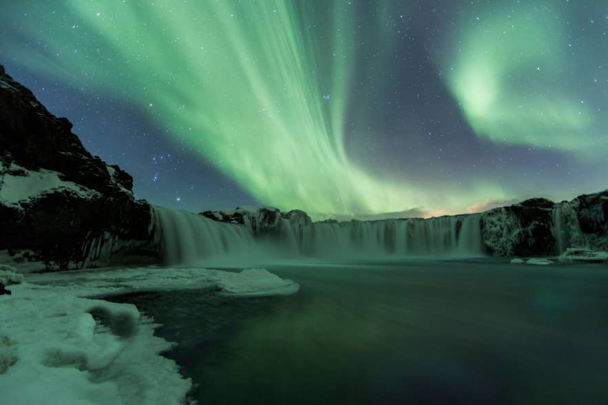 Aurora borealis over a frozen waterfall in Iceland, with green northern lights reflecting on icy water at night.