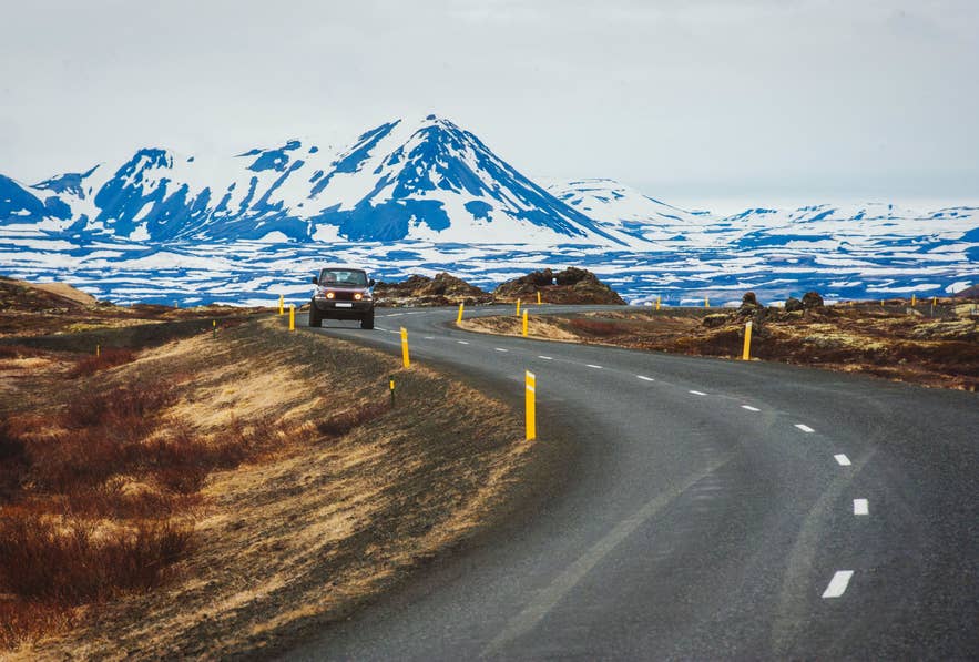 A rental car drives an open road toward snow-covered mountains in Iceland, where checking the wind forecast each morning decides the day's route in winter.