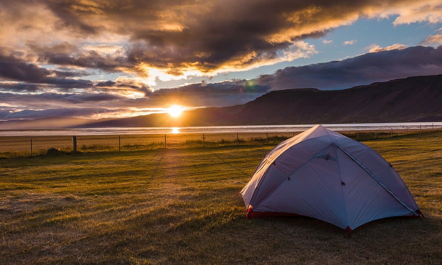 A camping tent at sunset by a fjord in Iceland in June, with golden skies, calm water, and mountains in the background.