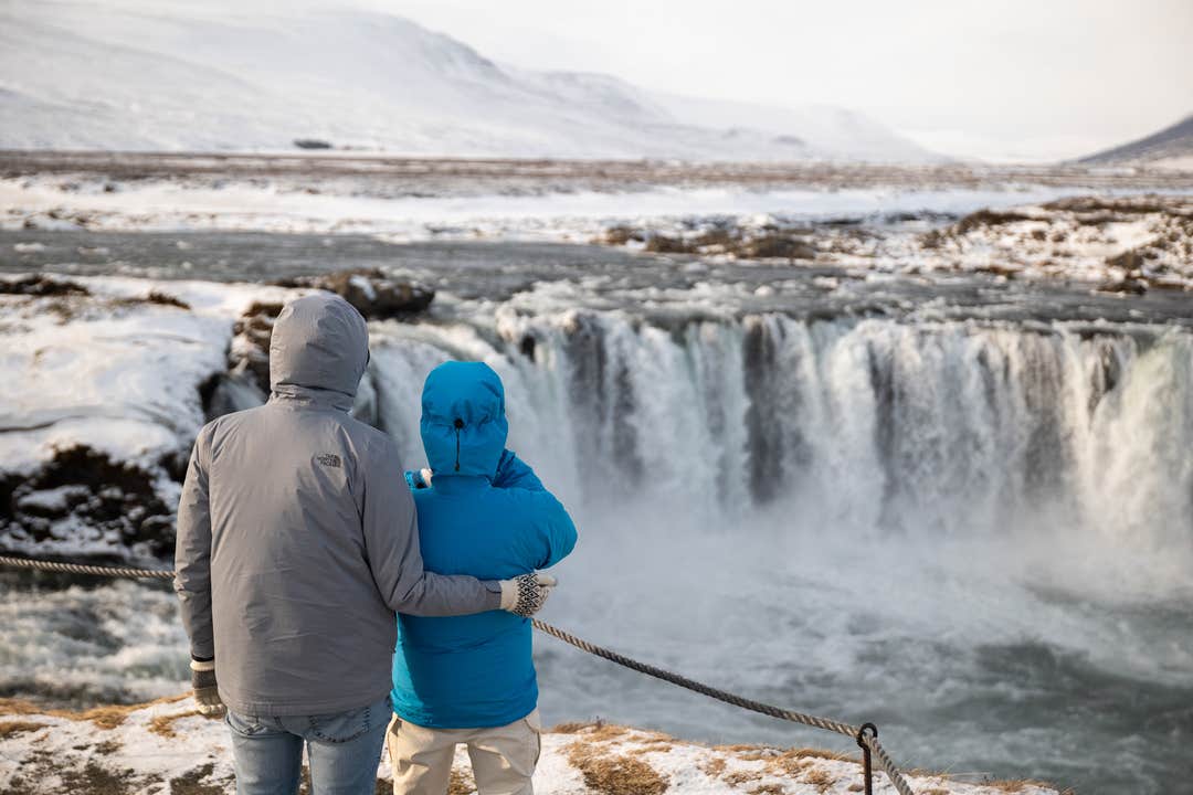 Zwei Touristen in Winterkleidung stehen mit dem Rücken zur Kamera und blicken auf die gefrorene Landschaft rund um den Godafoss-Wasserfall in Nordisland.