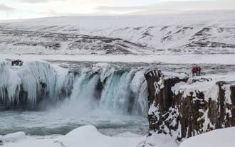 Un touriste lors d'une excursion à Godafoss se tient au sommet d'une falaise, mettant en valeur l'immensité de la chute d'eau de Godafoss.