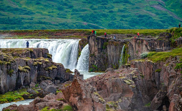 Besucher stehen auf Felsen am Godafoss-Wasserfall, wo mächtige Kaskaden durch Basaltformationen in Nordisland rauschen.
