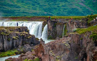 Besökare som står på klippiga avsatser vid Godafoss vattenfall, med kraftfulla kaskader som skär genom basaltformationer på norra Island.